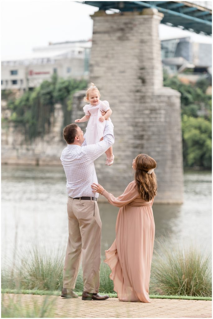 Mom in blush dress and dad in button down hold daughter in pale pink dress overhead with Tennessee River in the background during family maternity session in Chattanooga, TN. 