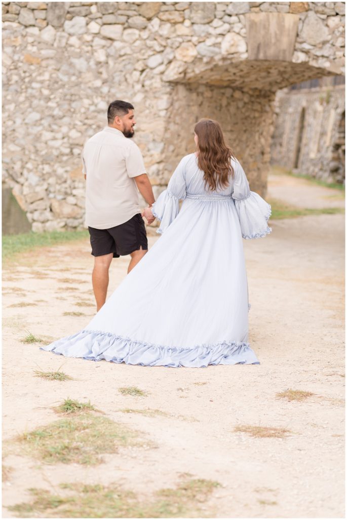 Couple hold hands and walk down path together with historic stone archway during maternity session in McKinney, TX.
