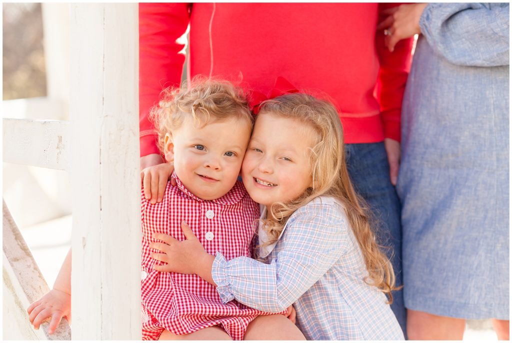 Brother and sister in red and blue gingham outfits smile and hug with mom and dad standing behind them during family session in Raleigh, NC with Wisp + Willow Photography Co. 