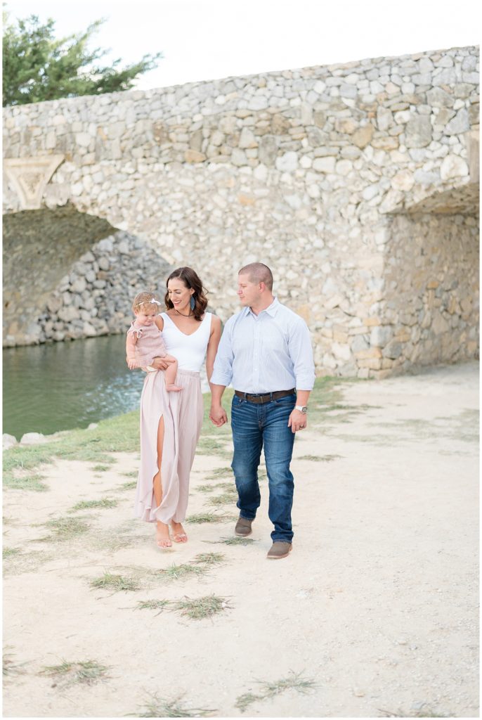 Mom in blush skirt and white tank holds hands and walks with husband in stripped button down shirt and jeans while holding baby daughter in mauve knit outfit with river and stone bridge in the background in Adriatica Village during family session.