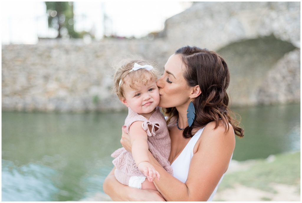 Mom in white tank and black feather earrings holds and kisses baby daughter in white bow and mauve knit outfit with river and stone bridge in the background during family photo session with Wisp + Willow Photography Co.
