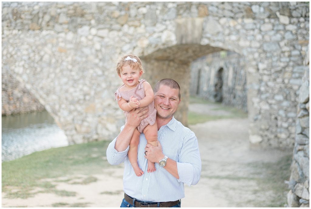 Dad in stripped button down holds baby daughter in mauve knit outfit on shoulder with historic stone bridge behind in Adriatica Village during family photo session.