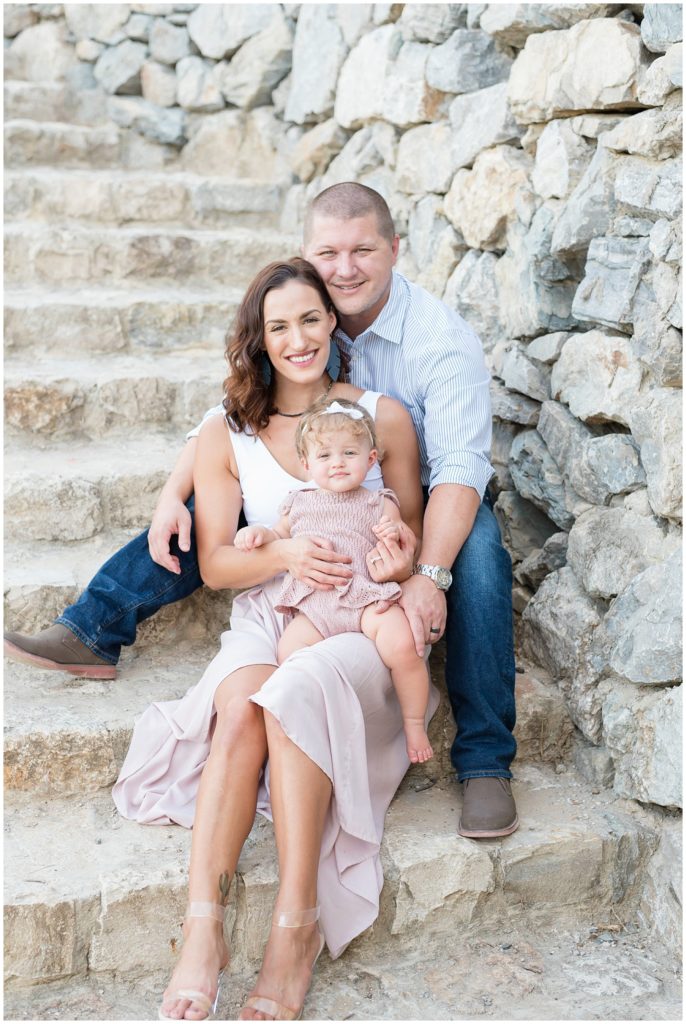 Mom in blush skirt and white dress and dad in stripped button down and jeans sit on stone steps with daughter in mauve knit outfit during first birthday session in McKinney, TX.
