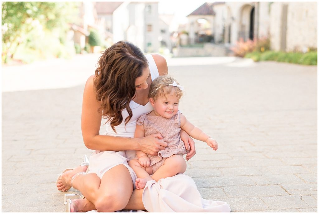 Mom in white tank and blush skirt sits on brick pathway in Adriatica Village with baby daughter on lap during family photo session with Wisp + Willow Photography Co.