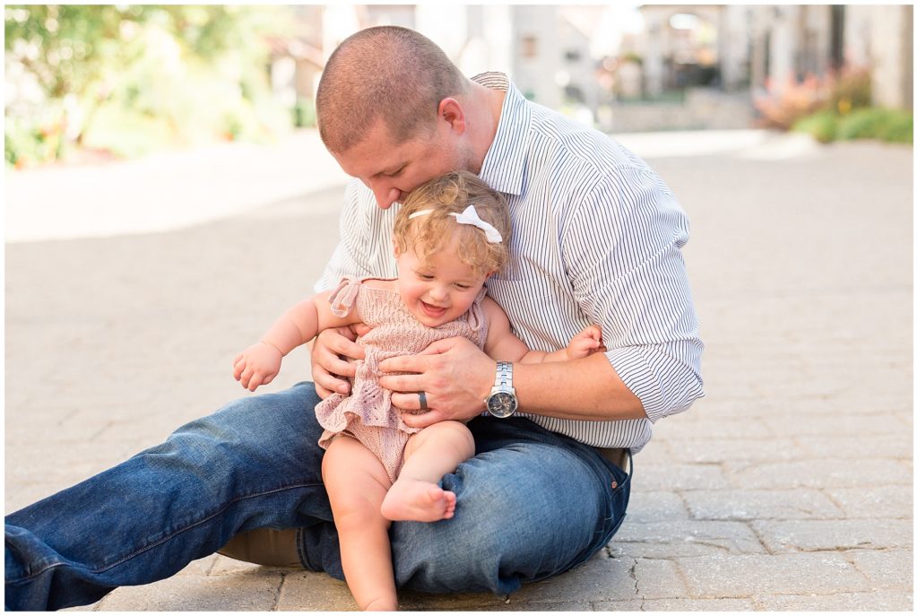 Dad in stripped button down and jeans sits and plays with baby daughter in mauve dress on brick pathway in McKinney, TX during family photo session.