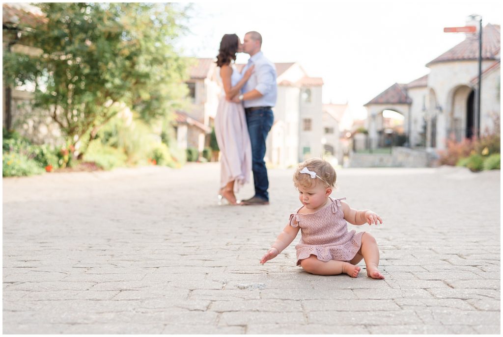 Mom and Dad kiss on brick pathway in Adriatica Village while one year old baby girl in mauve dress sits in front of them playing on the ground. 