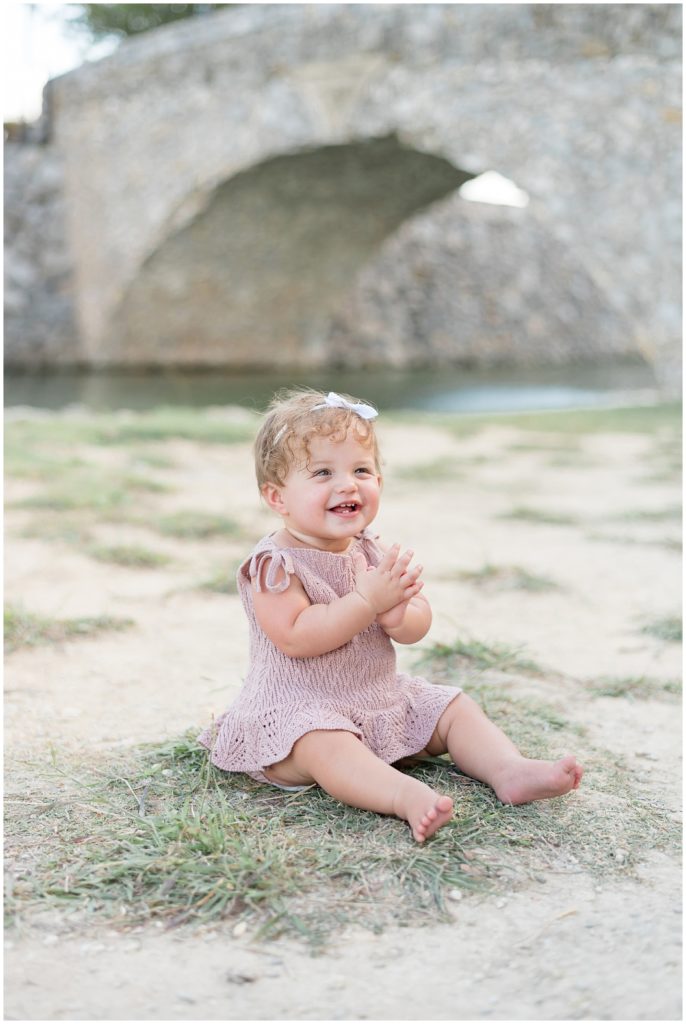 Baby girl in knit mauve dress and white bow sits in grass clapping with stone bridge behind during first birthday session with Wisp + Willow Photography Co. 