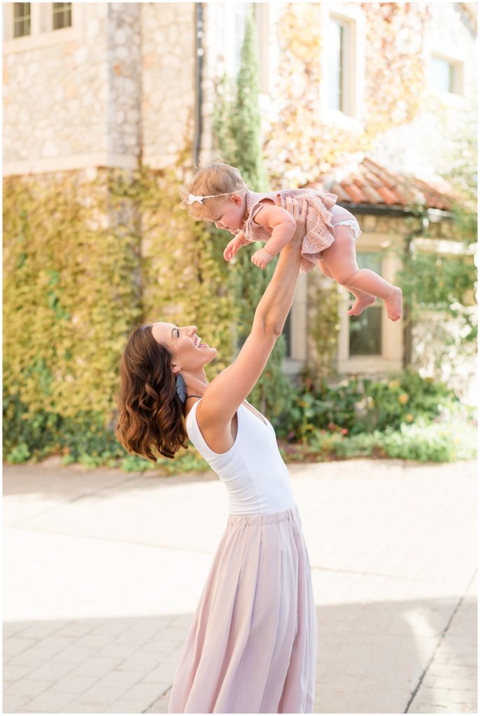 Mom in blush skirt and white tank holds baby in mauve dress overhead with historic house covered in vines behind during first birthday session in Adriatica Village. 