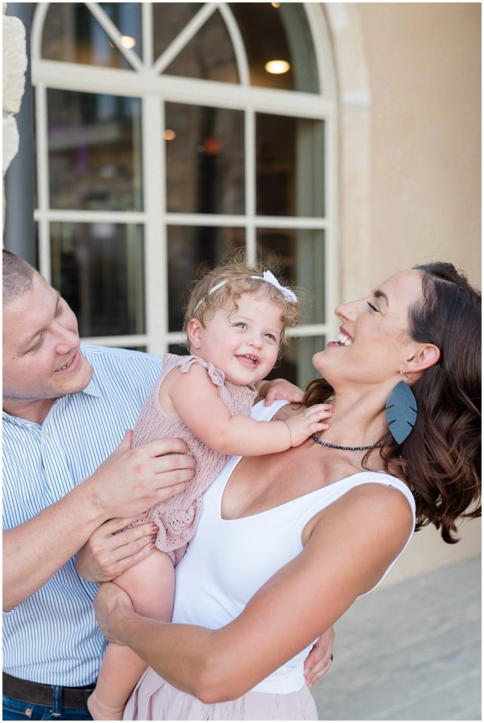 Mom in white tank and black feather earrings and Dad in stripped button down hold baby daughter in knit mauve outfit while tickling and laughing during firth birthday session in McKinney, TX. 