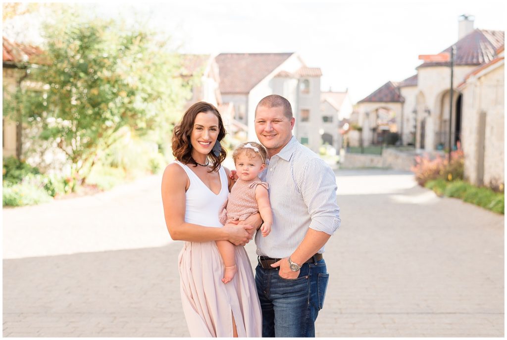 Mom in white tank and blush skirt and dad in button down and jeans hold daughter in mauve knit outfit on brick pathway in Adriatica Village during family photo session with Wisp + Willow Photography Co. 