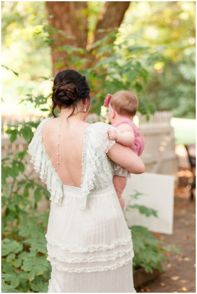 new mom in stunning free people white lace dress with open back holds 7 month old baby in pink chiffon romper and pink flower bow in historic oak view park in raleigh, north carolina taken by Wisp + Willow Photography Co
