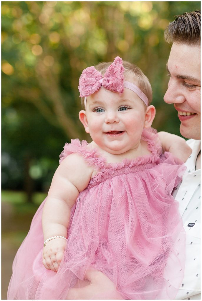 new dad holds 7 month old baby girl in pink dress and bow headband with a pearl bracelet around her wrist.  Click more to see this happy baby that smiles for the Wisp + Willow Photography Co.