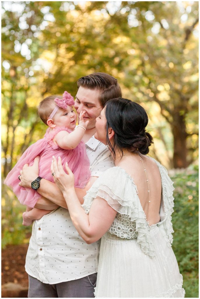 family of 3 hugs in close as 7 month old baby girl is held by dad and reaches for mom.  gorgeous free people white dress worn by mom and pink dress and headband work by baby girl and dad wears white shirt with blue polka dots in garden.