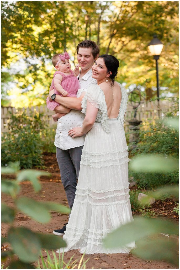 new mom wears gorgeous open back free people white dress in garden while husband holding 7 month old daughter look at her.