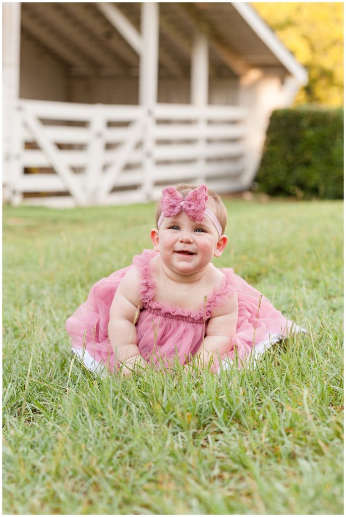 happy 7 month old baby girl in the grass at historic oak view park in raleigh north carolina wearing a pink chiffon dress and headband with a bow taken by Wisp + Willow Photography Co.