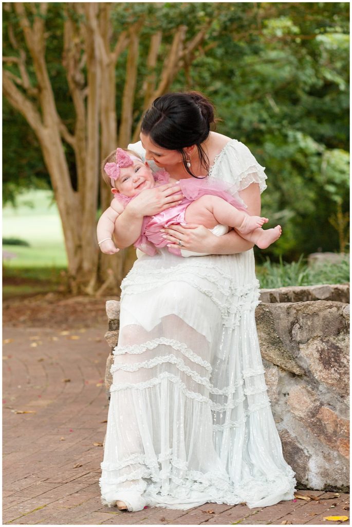 New mom holds 7 month old baby girl in family session taken in raleigh north carolina by Wisp + Willow Photography Co.  Mom is wearing a white free people dress and daughter wears a pink dress with pink headband and pearl bracelet.