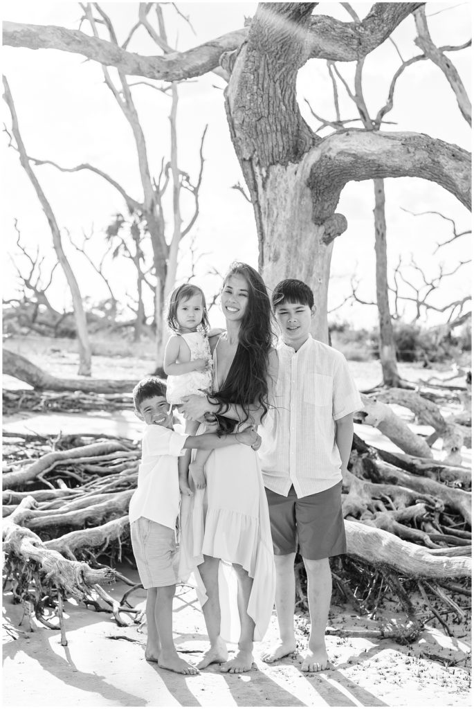 Mom stands in front of large pieces of driftwood on beach with 3 children in Jekyll Island, GA during Mommy & Me photo session with Wisp + Willow Family Photography Team. 
