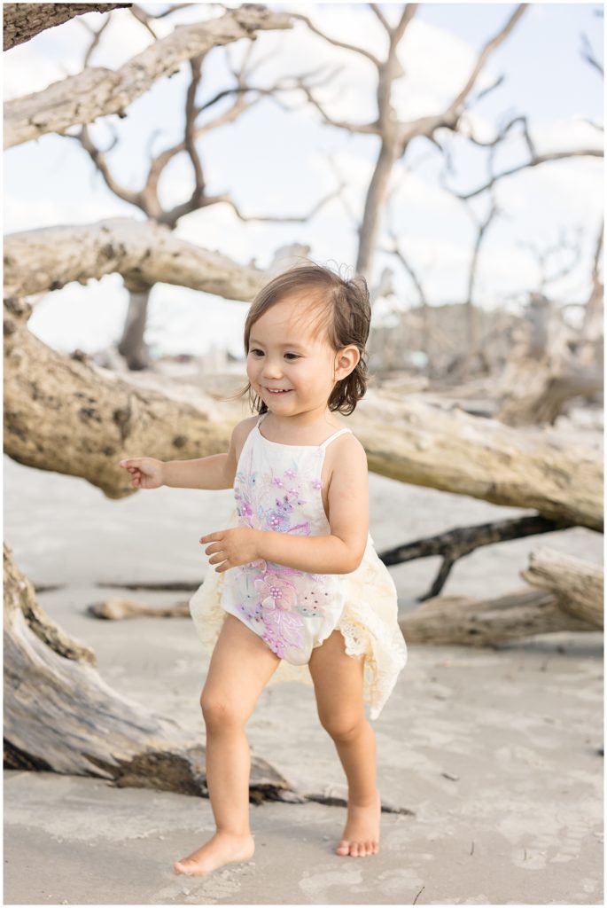 Little girl in white lacey outfit with purple flowers runs and plays on beach with driftwood behind her during family photo session in Jekyll Island, GA. 