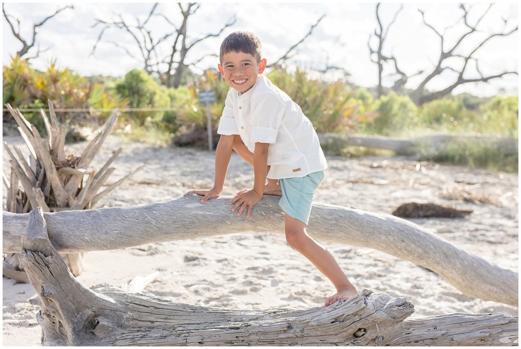 Boy in white button down shirt and green shorts climbs on piece of driftwood on beach with greenery in the background during family photo session in Jekyll Island, GA. 