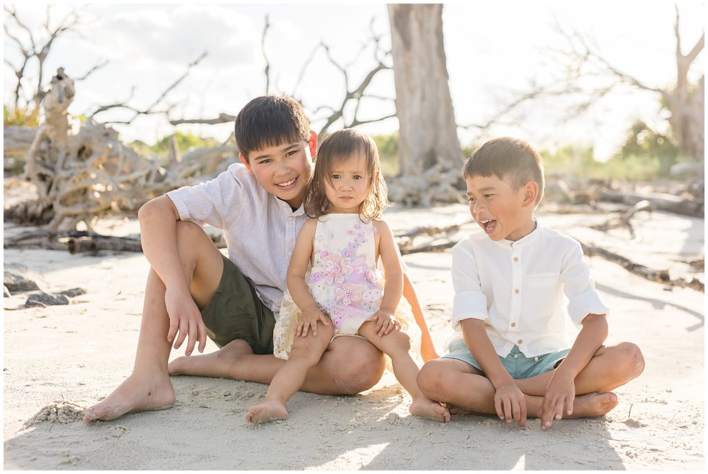 Two brothers in white button down shirts and green shorts and sister in white outfit with purple flowers sit in sand and laugh together at Driftwood Beach.