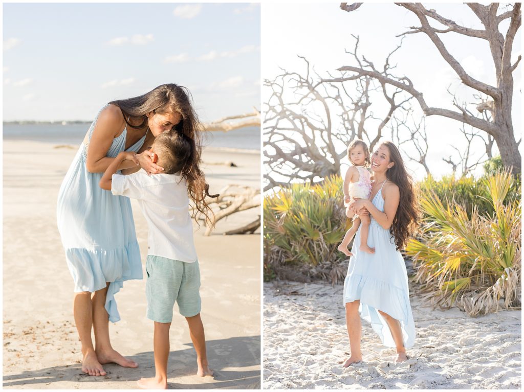 Mom in blue flowy dress kisses son in white button down and green shorts on forehead on Driftwood Beach. Mom in blue dress holds toddler daughter in white outfit in front of palm leaves and driftwood in Jekyll Island, GA. 