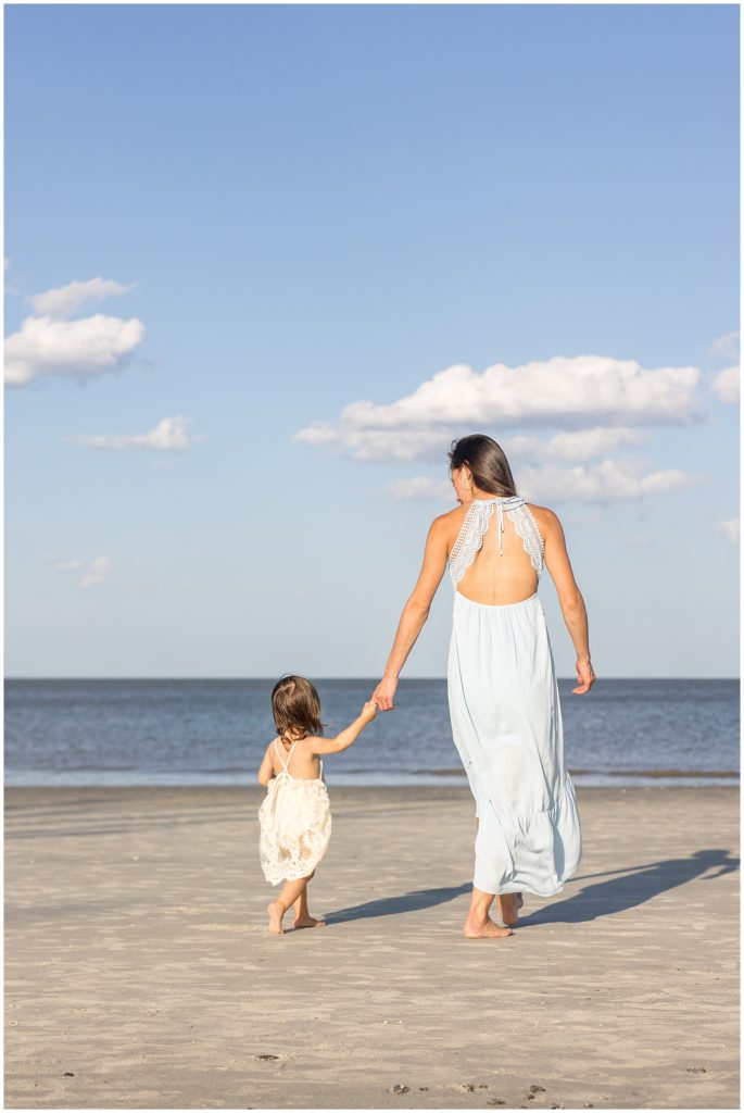 Mom in blue dress with lacey back holds toddler daughter's hand in white lacey dress while walking on Driftwood Beach during Mommy & Me photo session with Wisp + Willow Photography Co. 