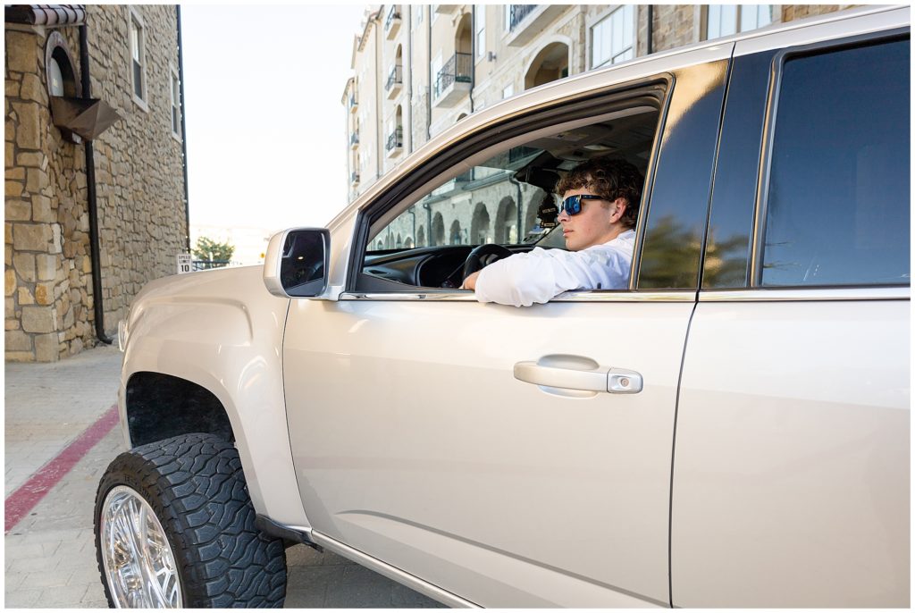 High school senior boy in white shirt and sunglasses sits in his silver truck while looking out at Adriatica Village during senior portrait session. 