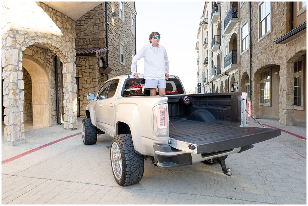Senior boy in white long sleeve shirt and grey shorts stand in the bed of silver pickup truck on cobblestone pathway between historic stone buildings. 
