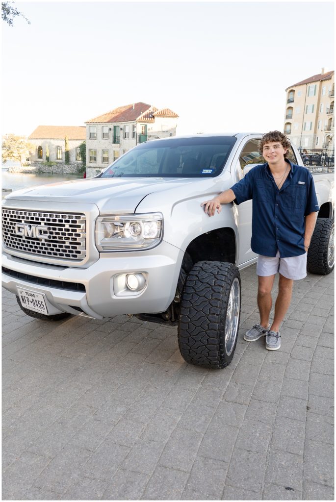 During senior photo session, high school boy in dark blue button down and grey shorts leans on silver pickup truck in McKinney, TX. 