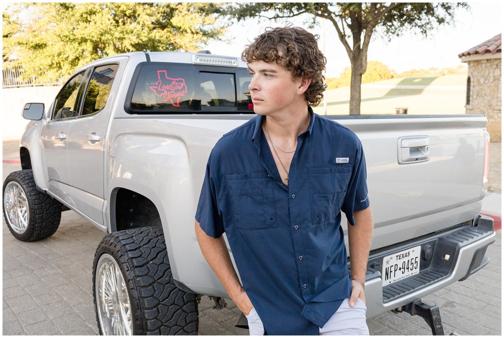 High school senior in dark blue button down and silver necklace leans on bumper on silver pickup truck during senior photo session in McKinney, TX. 