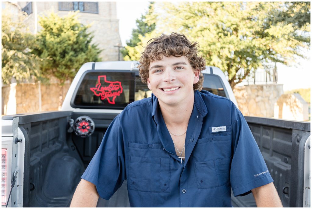 During senior portrait session, teenage boy in dark blue button down and silver necklace sits in bed of silver pickup truck with red Texas bumper sticker in McKinney, TX. 