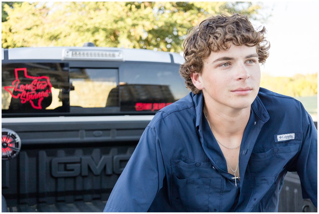 High school senior boy in dark blue button down and silver necklace sits in bed of silver pickup truck during senior portraits. 