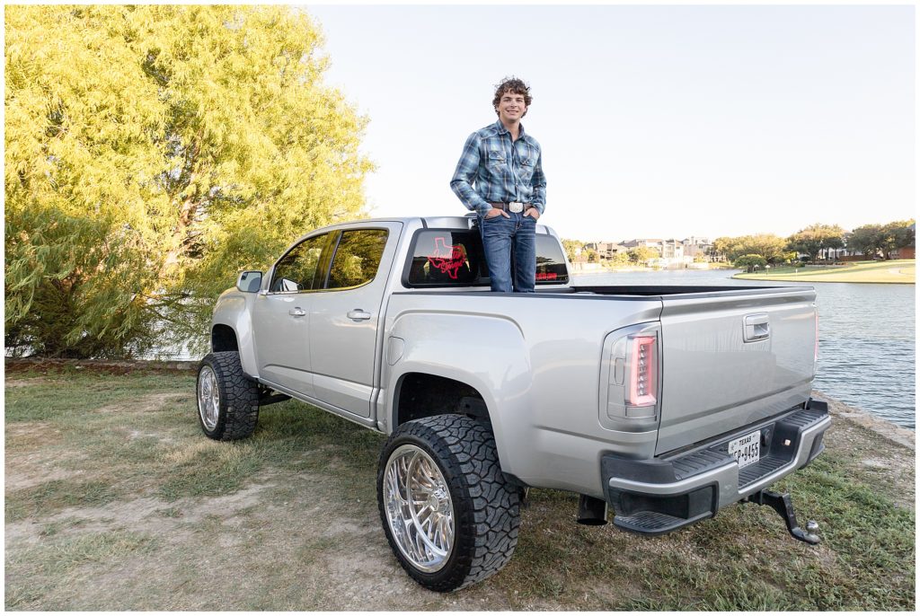 During senior photo session in McKinney, TX, teenage boy in blue plaid button down and jeans stands in the bed of silver pick up truck. 