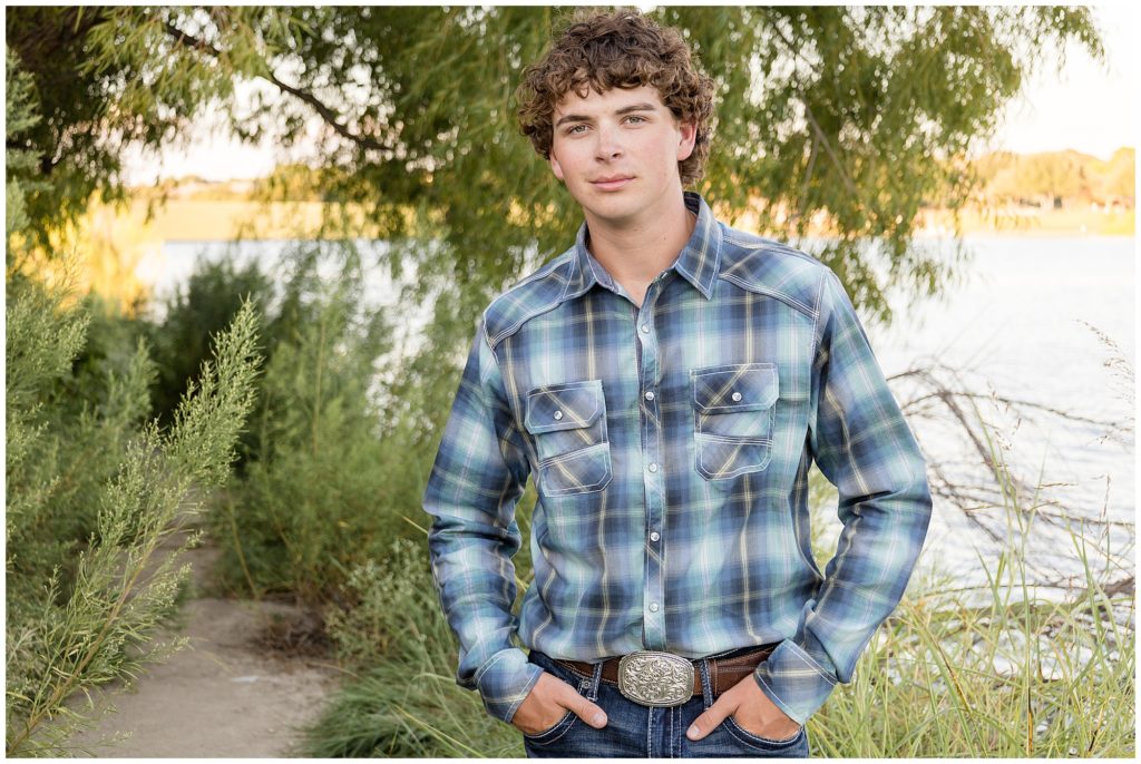 High school senior boy in blue plaid button down, jeans, and belt stands in front of lake and trees during senior photo session with Wisp + Willow Photography Co. 