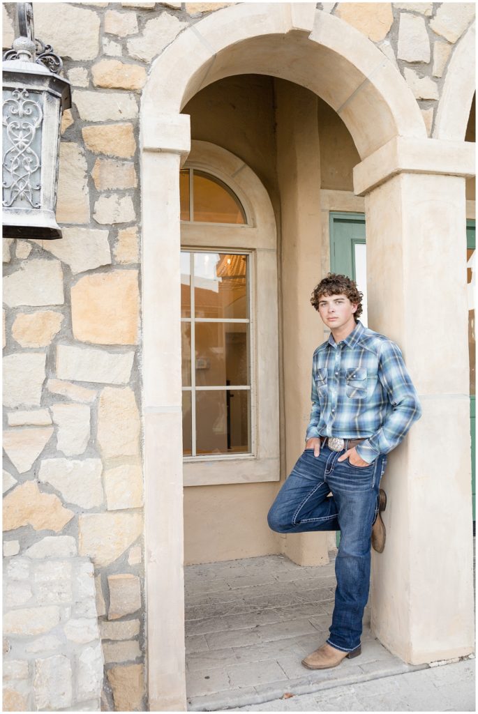 High school senior in blue plaid button down, jeans, and cowboy boots leans on stone doorway in Adriatica Village during senior photo session. 