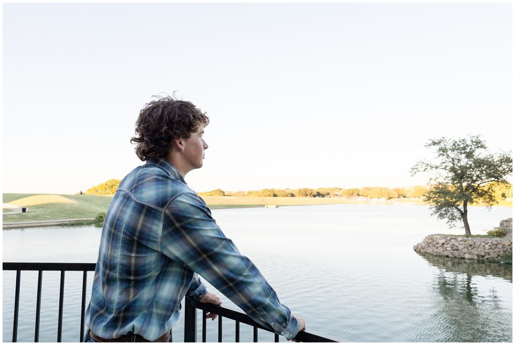 High school senior in blue plaid button down leans on railing while looking out over lake in Adriatica Village in McKinney, TX. 