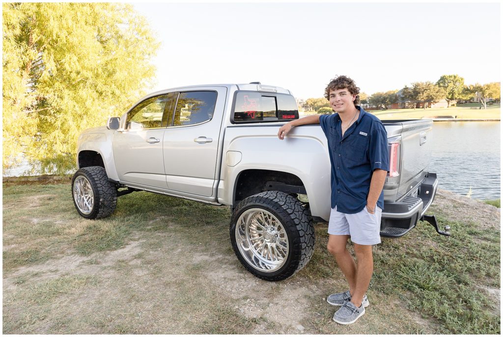Senior boy in dark blue button down and grey shorts leans on silver pickup truck in Adriatica Village during senior photo session. 