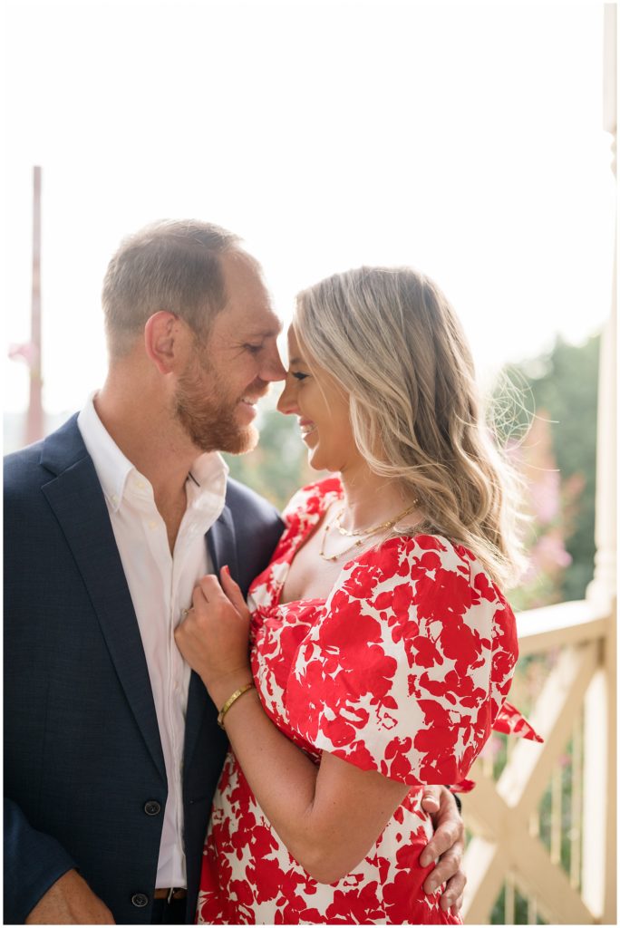 Man in blue suit holds fiance in red and white dress with gold jewelry close on white balcony during engagement session in Libby Hill. 