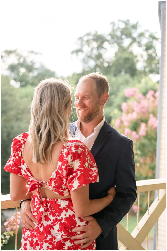 Woman in red and white dress holds fiance in blue suit close while standing on white balcony with flowers in the distant during engagement session with Wisp + Willow Photography Co. 