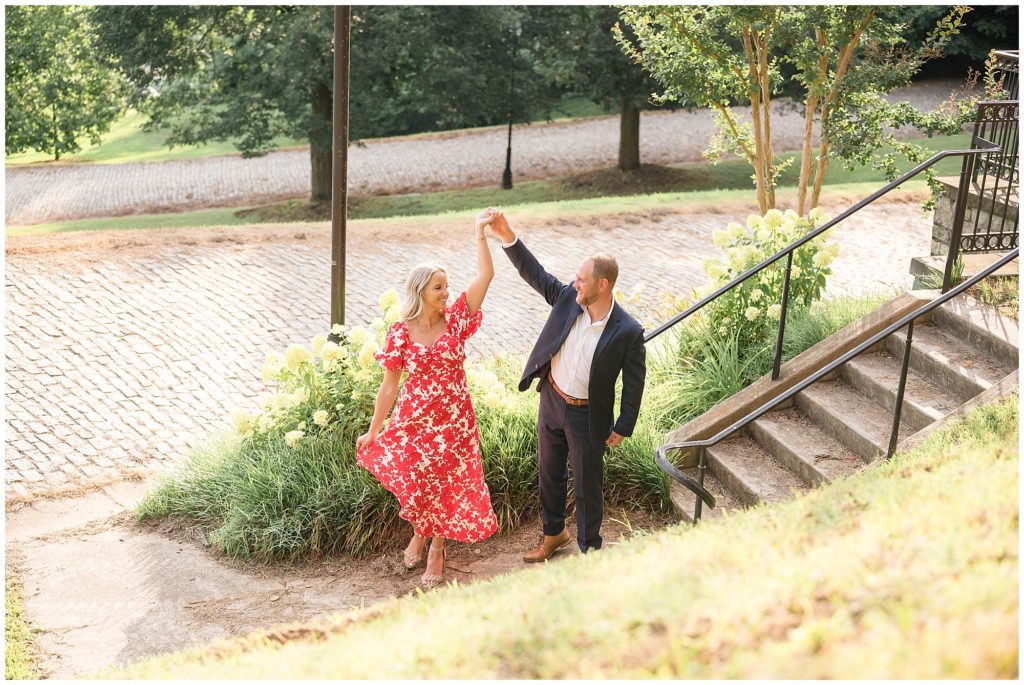 Engaged couple in red and white dress and blue suit dance on pathway in Libby Hill park with cobblestone pathways and trees around. 