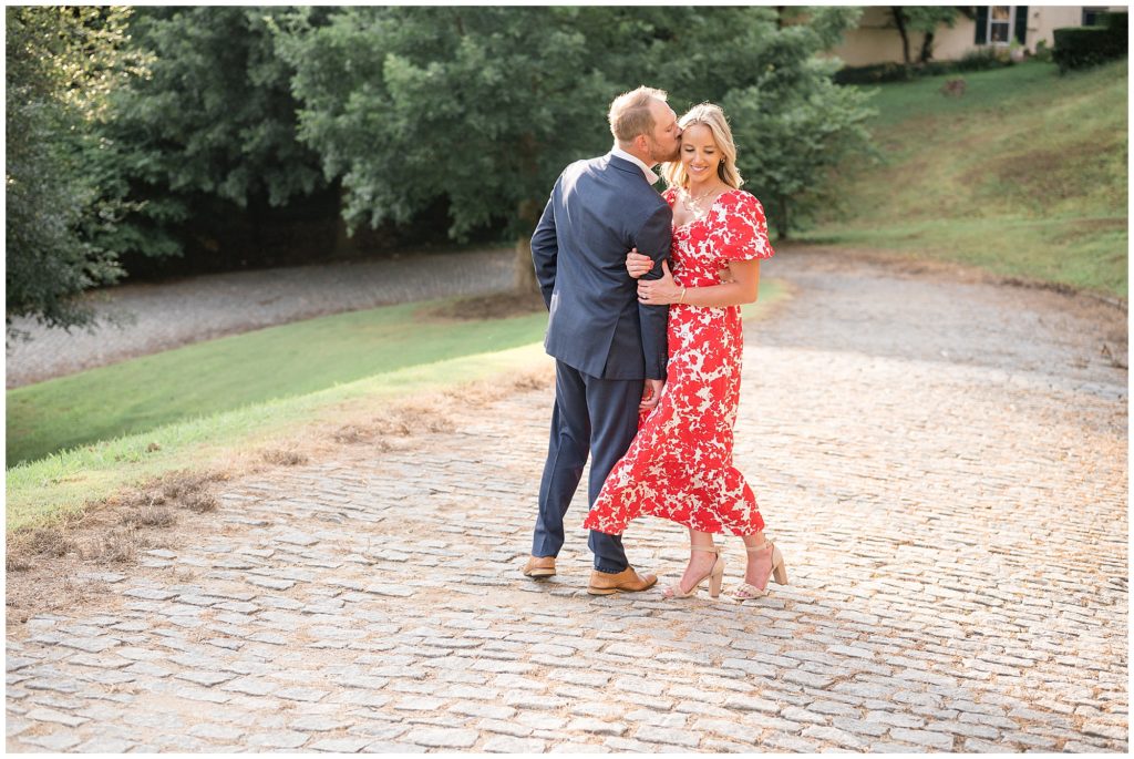 Man in blue suit leans over and kisses fiance in red and white dress on forehead while standing on cobblestone pathway in Richmond, VA. 