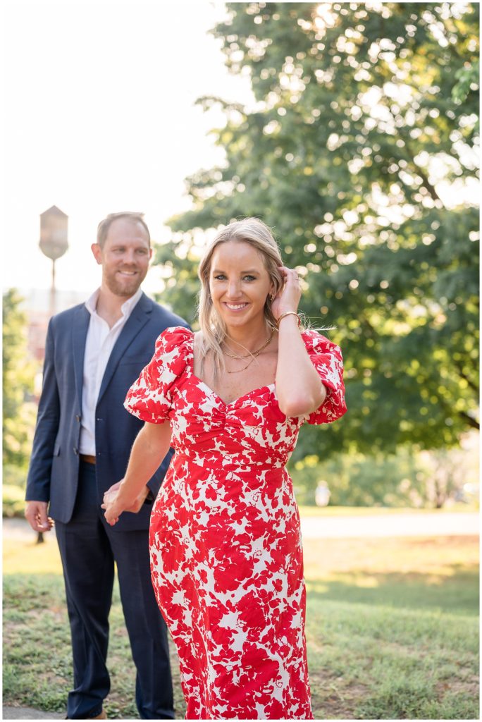 Woman in red and white dress and gold jewelry holds hands with fiance in blue suit while walking through grass in Libby Hill in Virginia. 