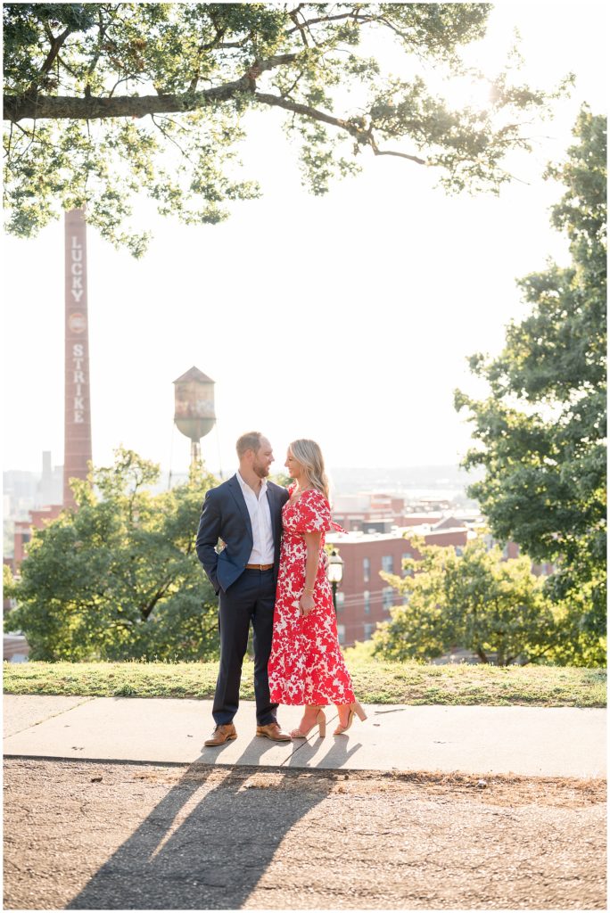 Couple in blue suit and red and white dress stand in on sidewalk in Libby Hill overlooking Richmond, VA during engagement session. 