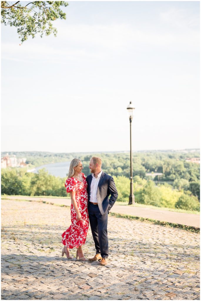 Beautiful engaged couple in red and white dress and blue suit stand on cobblestone pathway overlooking river and forest in Richmond, VA. 