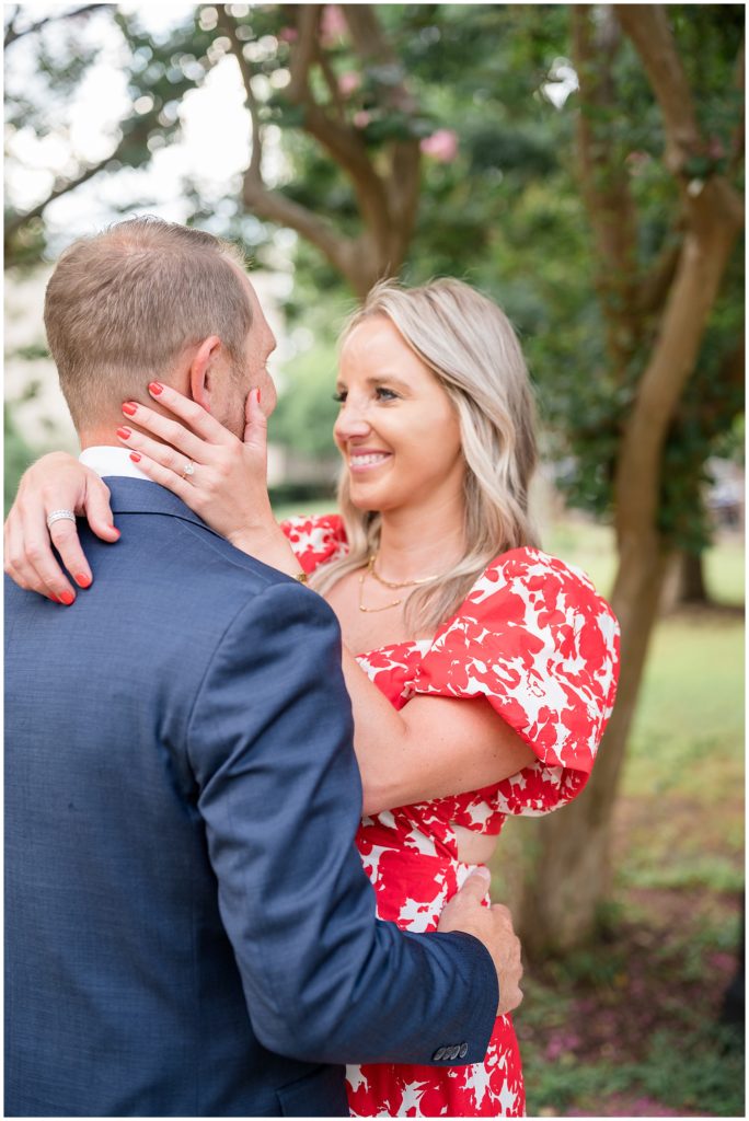 Woman in red and white dress with gold jewelry wraps arms around fiance in blue suit during engagement session with Wisp + Willow Photography Co. 