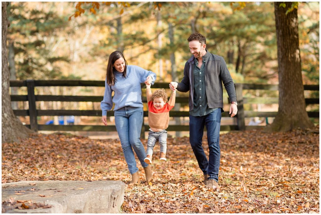 Mom in blue sweater and jeans and dad in green button down and jeans swing toddler son in bed of autumn leaves in Suwanee, GA during fall family photo session. 