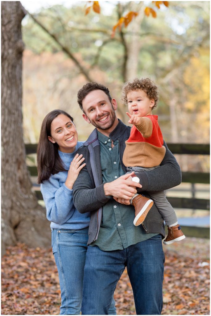 Mom and dad hold toddler son in red and brown sweater and smile with trees and black fence in the background during fall family photo session. 