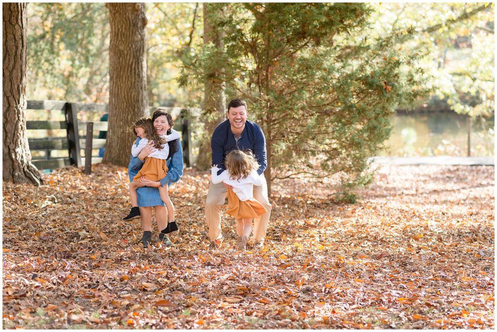2 sisters in orange and white outfits run and hug parents in bed of autumn leaves at Sims Lake Park in Suwanee, GA. 