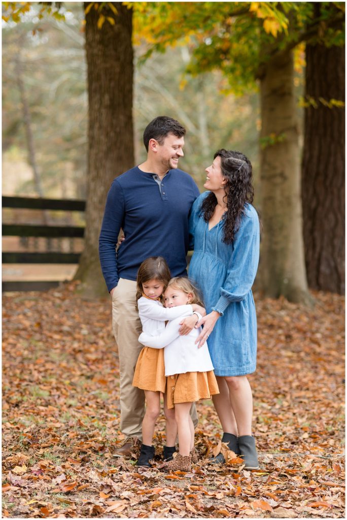 Mom in dad in blue dress and shirt stand behind 2 little daughters hugging in matching orange dresses and white sweaters in Suwanee, GA during fall family photo session. 