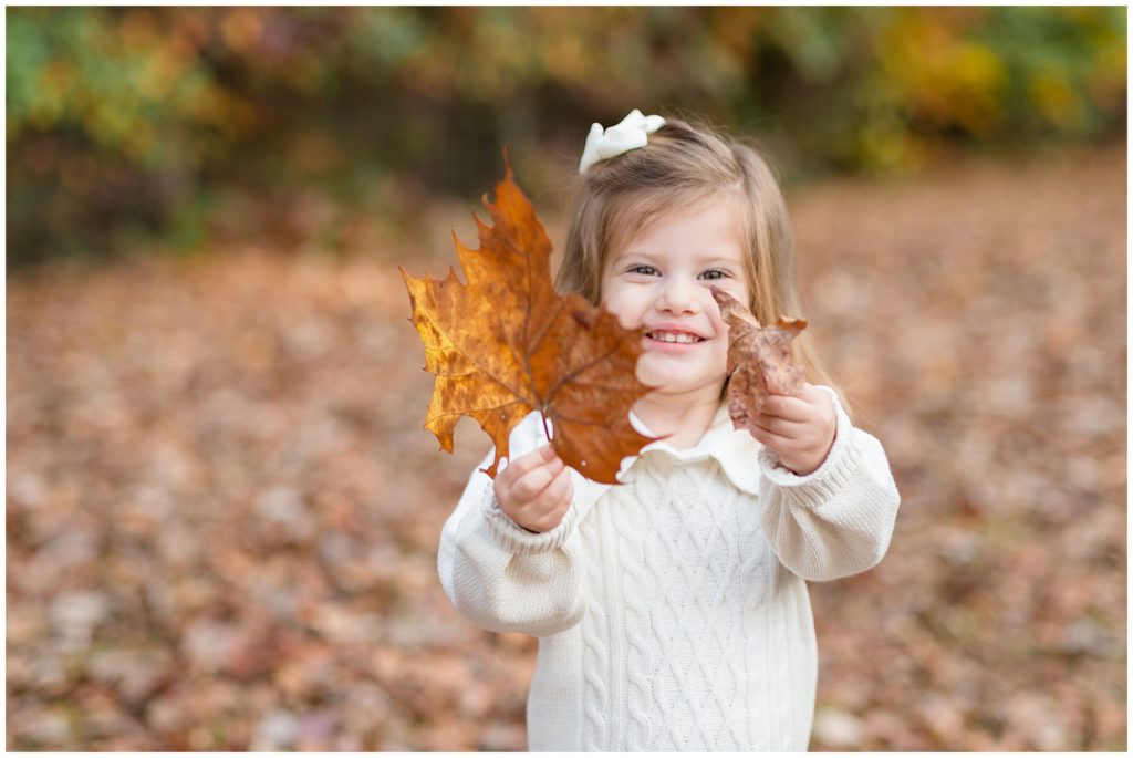 Little girl in white sweater and white bow smiles and holds large autumn leaves during fall family photo session in Suwanee, GA. 